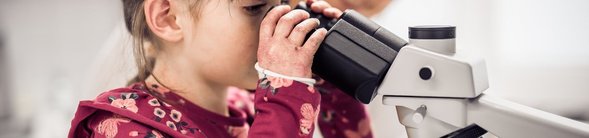 A little girl looks through a microscope with a researcher in the background