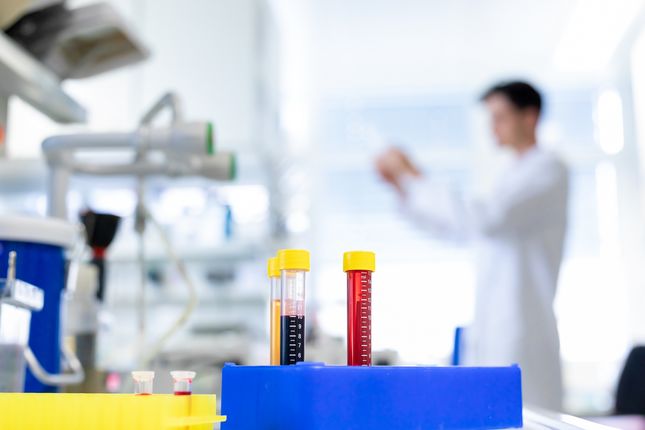 Laboratory situation: a researcher in the background and colourful pipettes in the foreground