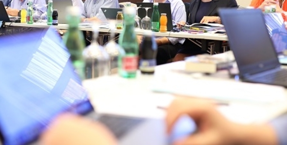 Conference situation with laptops in the foreground and row of people in the background.