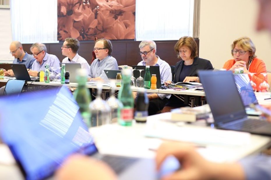 Conference situation with laptops in the foreground and row of people in the background.